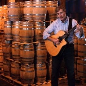 A man standing in front of wine barrels with an acoustic guitar.
