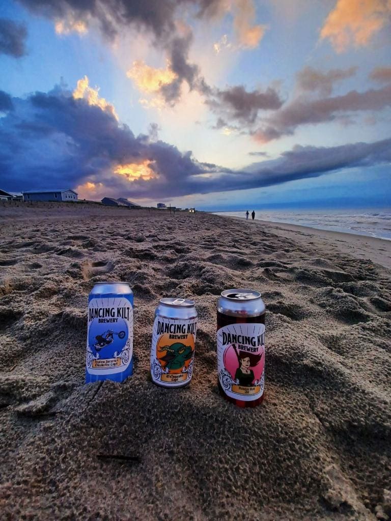 Three cans of beer sitting on the sand at sunset.