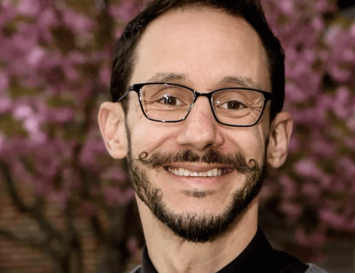 A man with glasses and a mustache smiles in front of flowers.