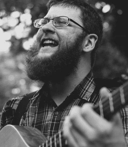 A man with glasses and a beard playing an acoustic guitar.